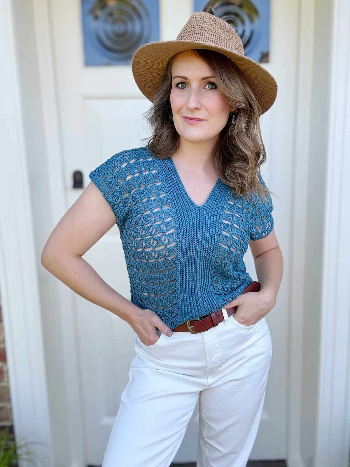 Person standing in front of a door, wearing a blue crocheted top, white pants, brown belt, and a straw hat.