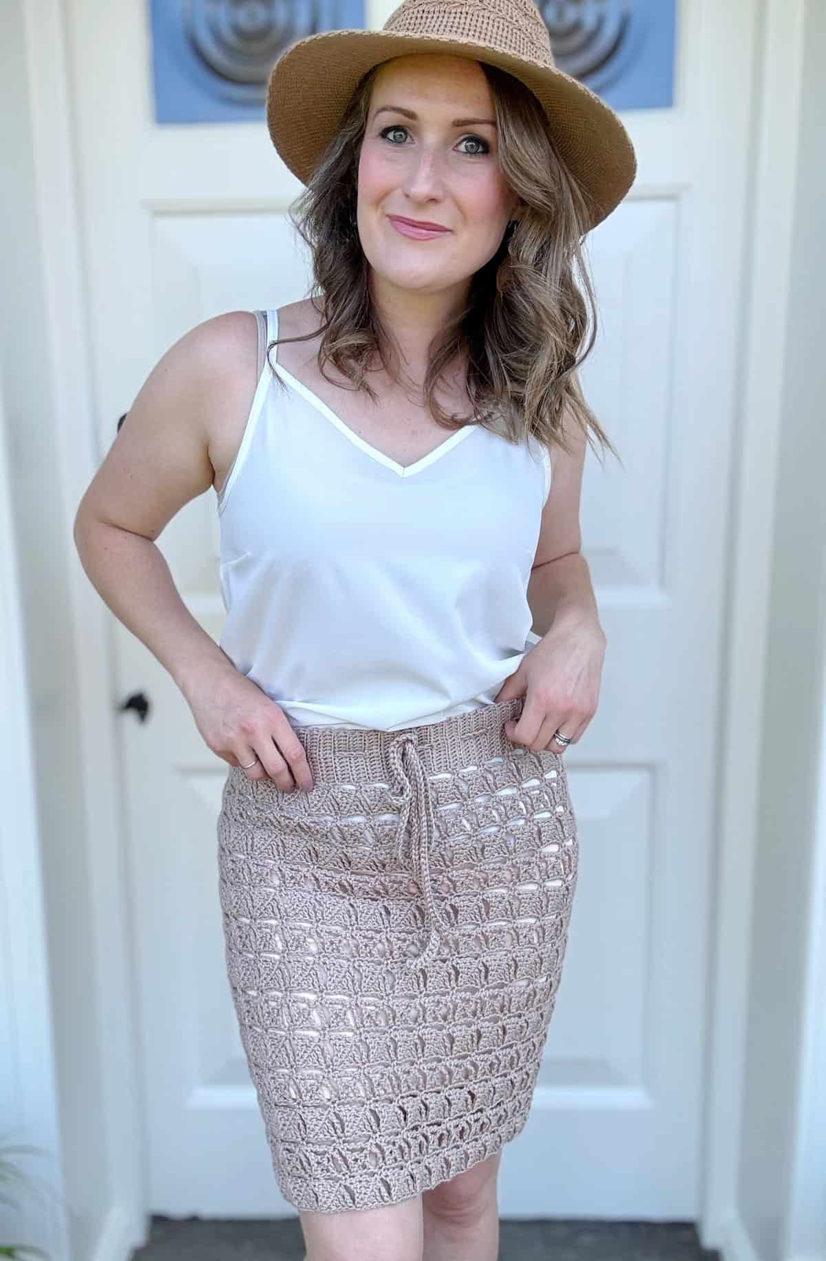 A woman stands in front of a white door wearing a white sleeveless top, a crochet skirt pattern in beige, and a woven broad-brim hat. She is smiling and has her hands on her hips.