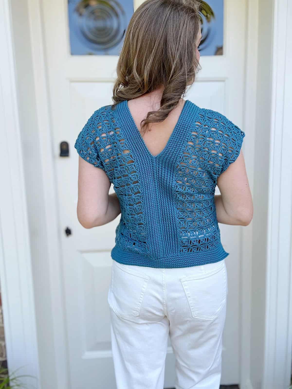 A person with long hair stands facing a white door, wearing a blue crochet lace top with a V-shaped back and short sleeves, paired with white pants.