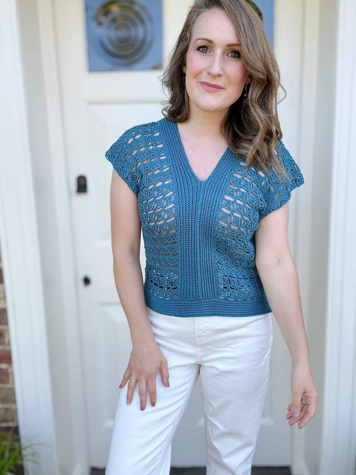A person wearing a blue crocheted top and white pants stands in front of a white door with a slight smile.