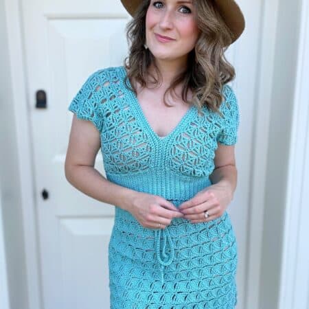 A woman wearing a light blue crochet skirt with top and a brown hat stands in front of a white door, looking at the camera with a slight smile.