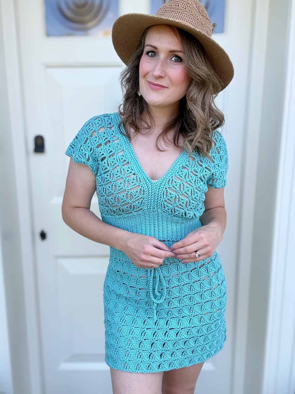 A woman wearing a light blue crochet skirt with top and a brown hat stands in front of a white door, looking at the camera with a slight smile.