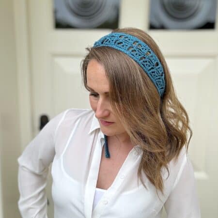 A woman with shoulder-length hair wears a white blouse and a blue crocheted headband, standing in front of a light-colored door.