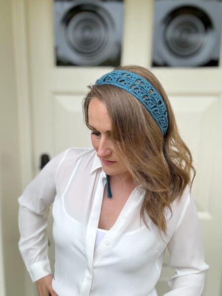 A woman with shoulder-length hair wears a white blouse and a blue crocheted headband, standing in front of a light-colored door.