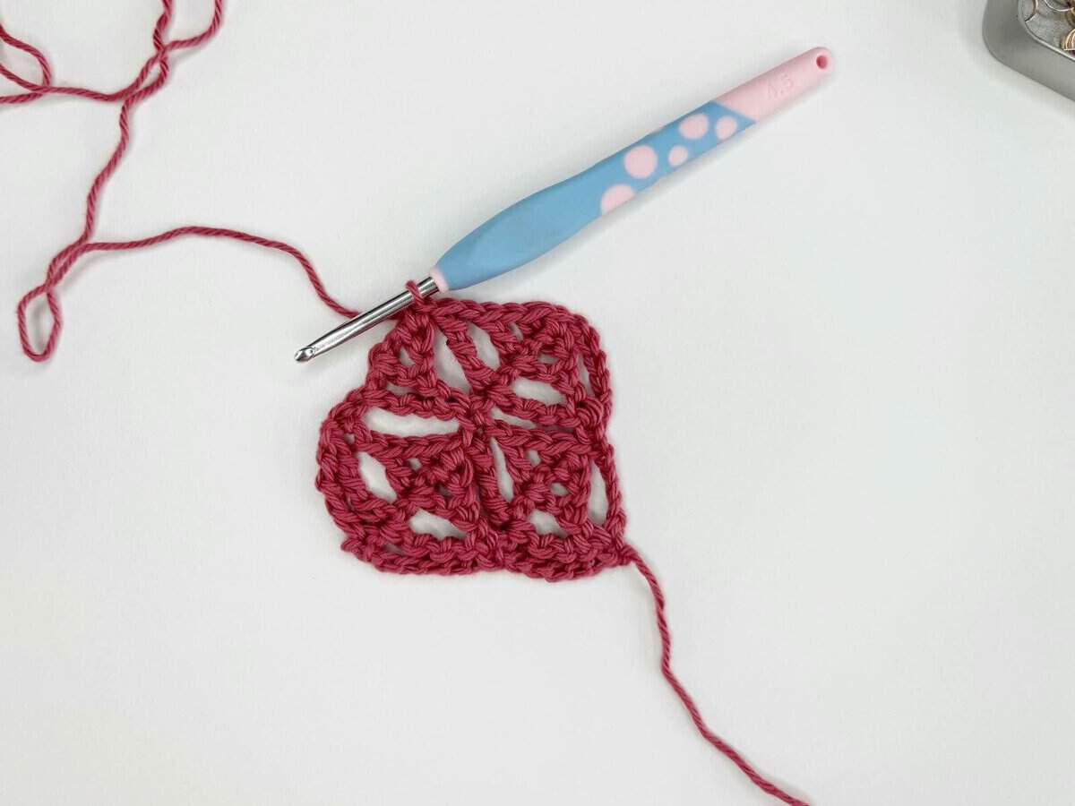 A partially completed heart-shaped crochet piece in red yarn, with a blue and pink crochet hook inserted into it, lies on a white surface.