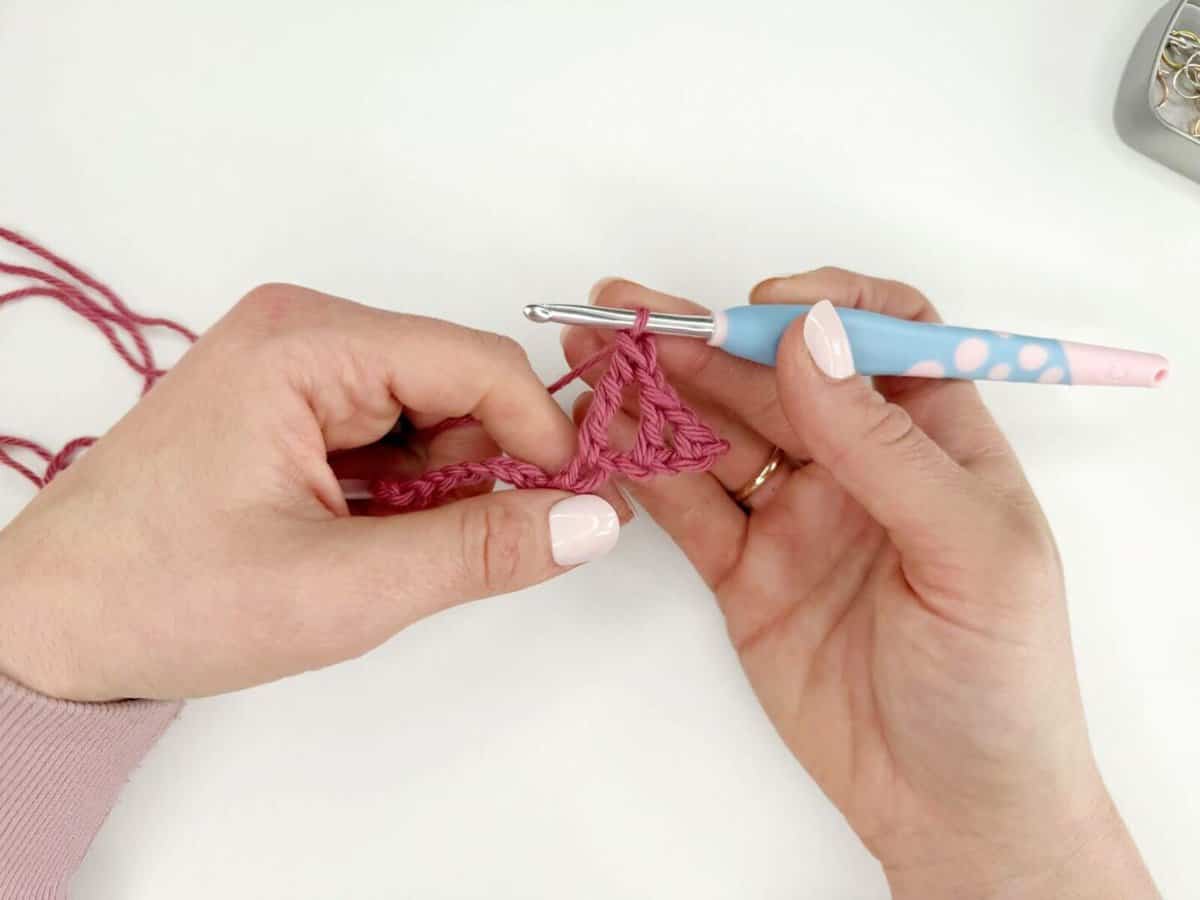 Close-up of hands holding a crochet hook and working pink yarn with long crochet stitches on a white surface.