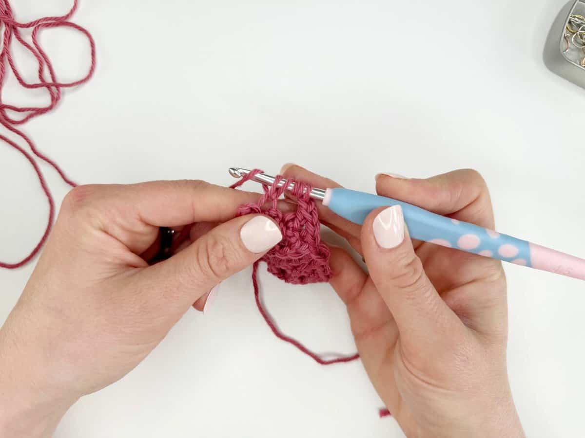 Hands crocheting long crochet stitches with pink yarn using a blue and pink crochet hook on a white surface. Loose yarn is visible in the background.