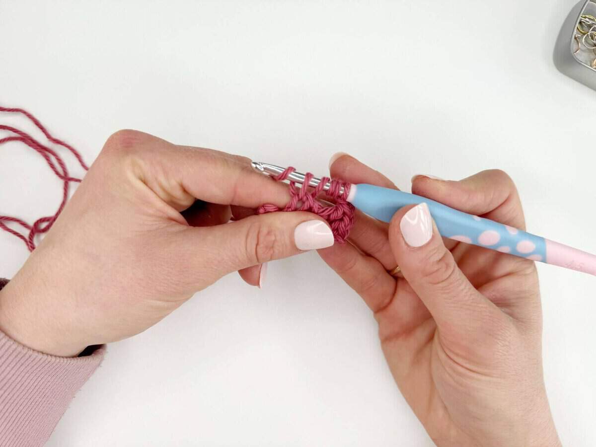 A person is using a pastel-colored crochet hook and pink yarn to create long crochet stitches. A small metal box with pins is partially visible in the upper right corner.