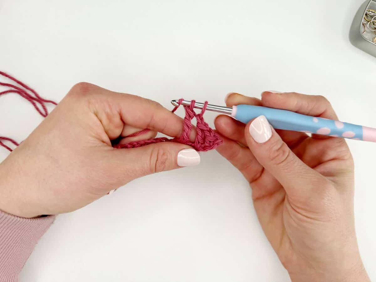 Close-up of hands crocheting with pink yarn and a blue crochet hook, showing long crochet stitches in progress against a white background.