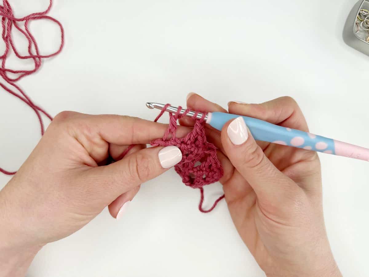 Two hands are crocheting a pink fabric with long crochet stitches, using a blue and pink crochet hook.