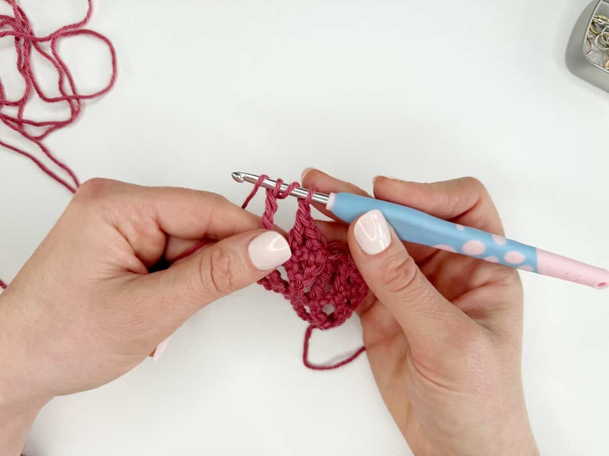 Close-up of a person crocheting with a blue and pink crochet hook and pink yarn, creating long crochet stitches against a white background.