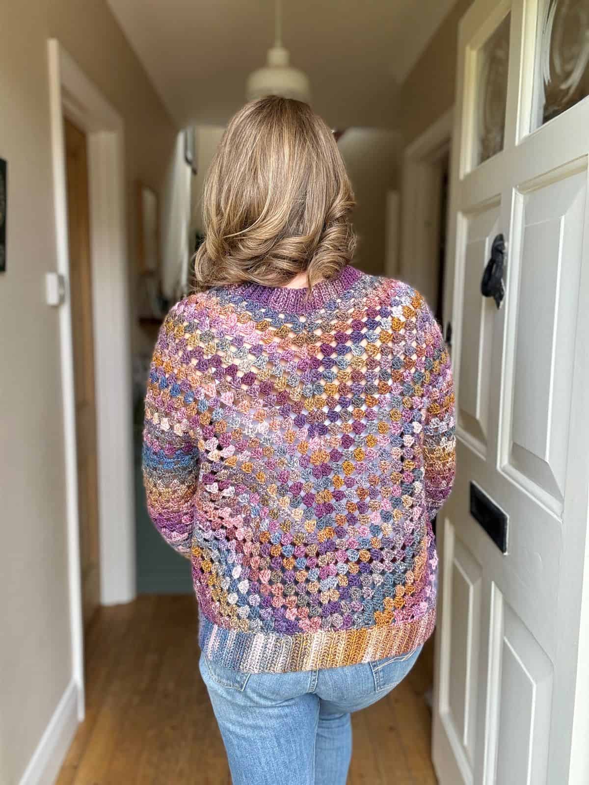 A person with shoulder-length hair, dressed in a colorful granny stitch crochet cardigan and jeans, is standing in a hallway with their back facing the camera.