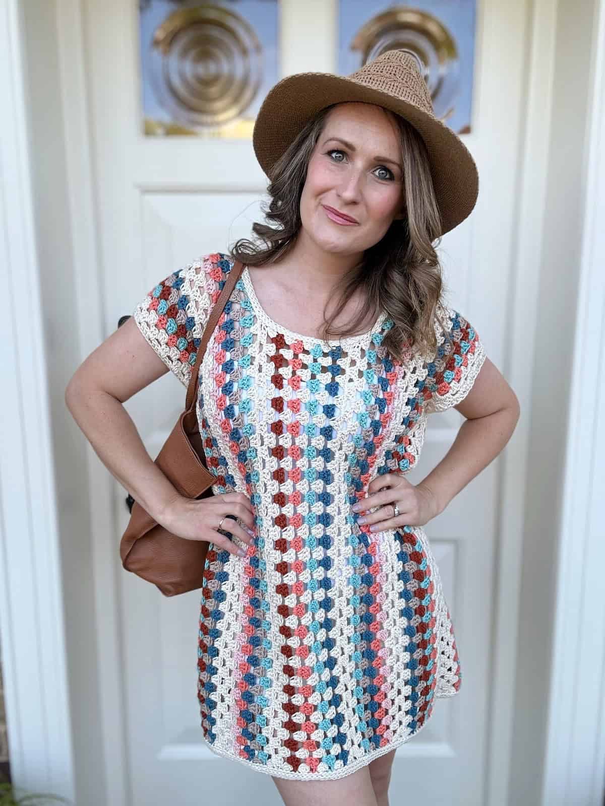 A woman stands in front of a white door, wearing a multicolored crochet dress with short sleeves and a wide-brimmed hat. She is looking at the camera with her hands on her hips and a brown purse over her shoulder.