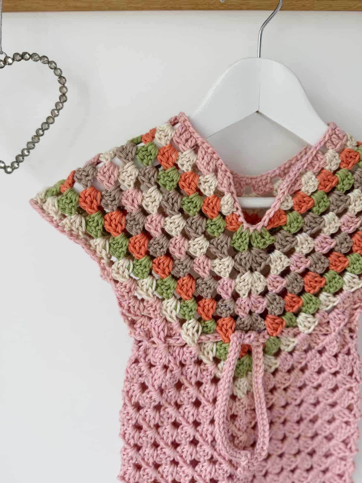 A crocheted garment with a diamond pattern in pink, green, white, and orange hangs on a white hanger next to a heart-shaped beaded decoration; it could easily inspire your next crochet dress pattern for baby.