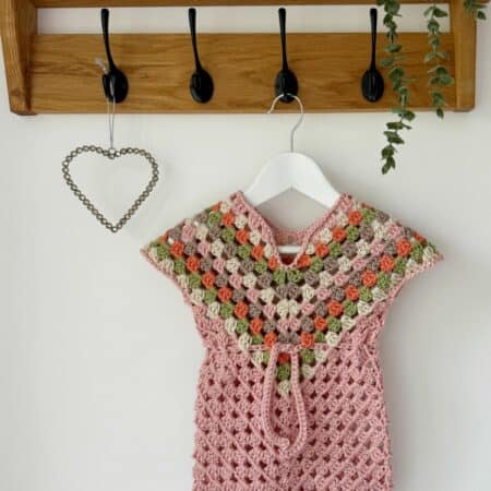 A crocheted pink dress with green and orange accents is hanging on a white hanger beneath a wooden rack with black hooks, next to a metal heart decoration and a small plant. This is an adorable crochet dress pattern for baby, adding charm to the cozy scene.