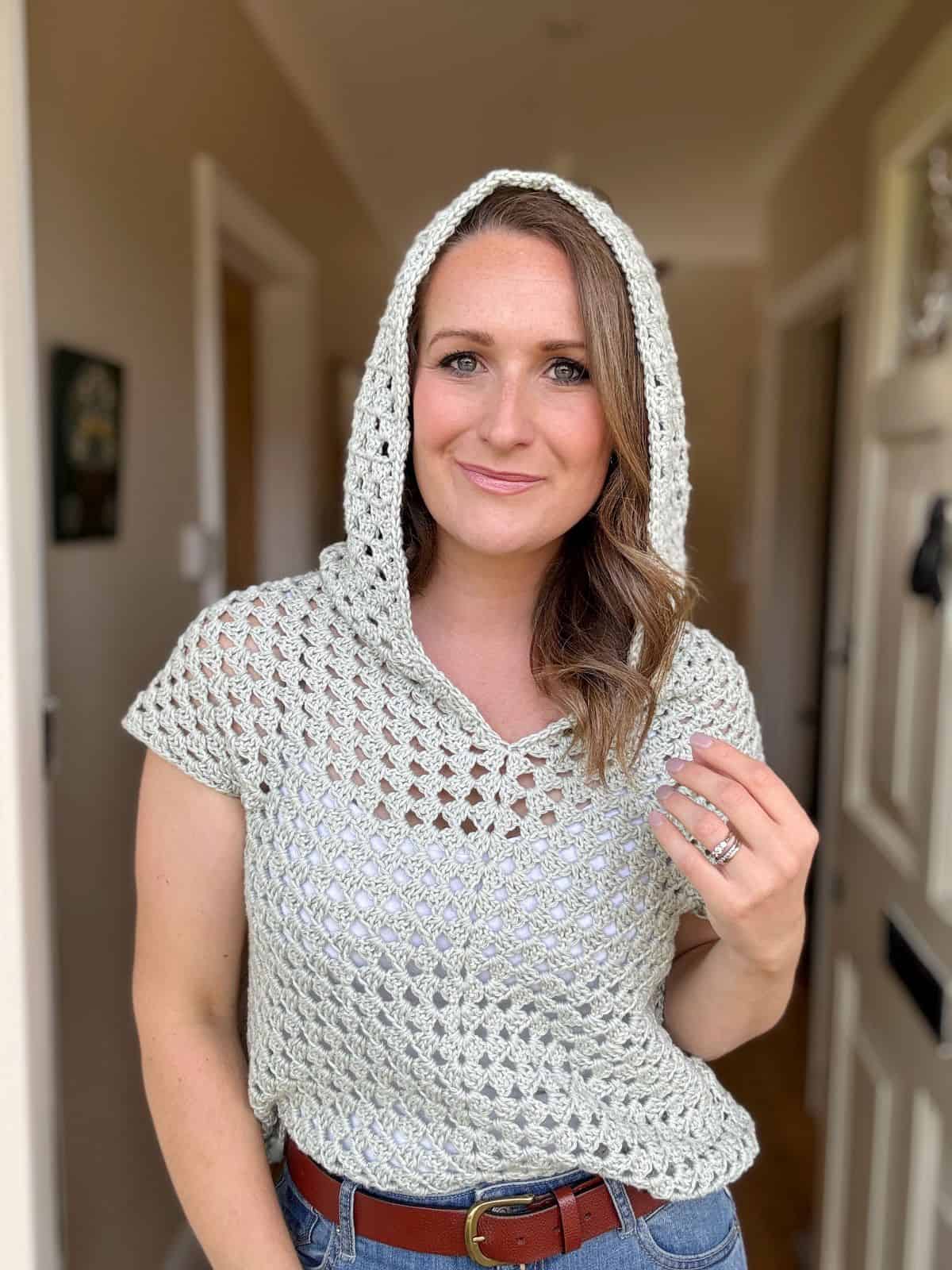 Person stands in a hallway wearing a light crocheted hooded top, showcasing an intricate crochet hoodie pattern, jeans, and a brown belt. They have shoulder-length wavy hair and are looking towards the camera with a neutral expression.