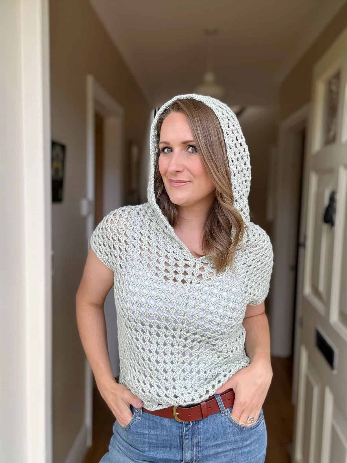 A woman standing in a hallway wears a light-colored, crocheted short-sleeve top with a hood, showcasing an intricate crochet hoodie pattern. Paired with jeans and a brown belt, she has her hands in her pockets and faces the camera.