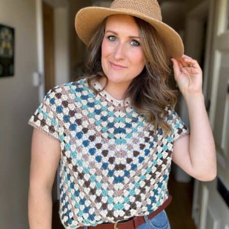 A woman is posing indoors, wearing a granny stitch crochet top, blue jeans, and a straw hat, touching the brim of the hat with her right hand. She has wavy hair, and there is a door in the background.