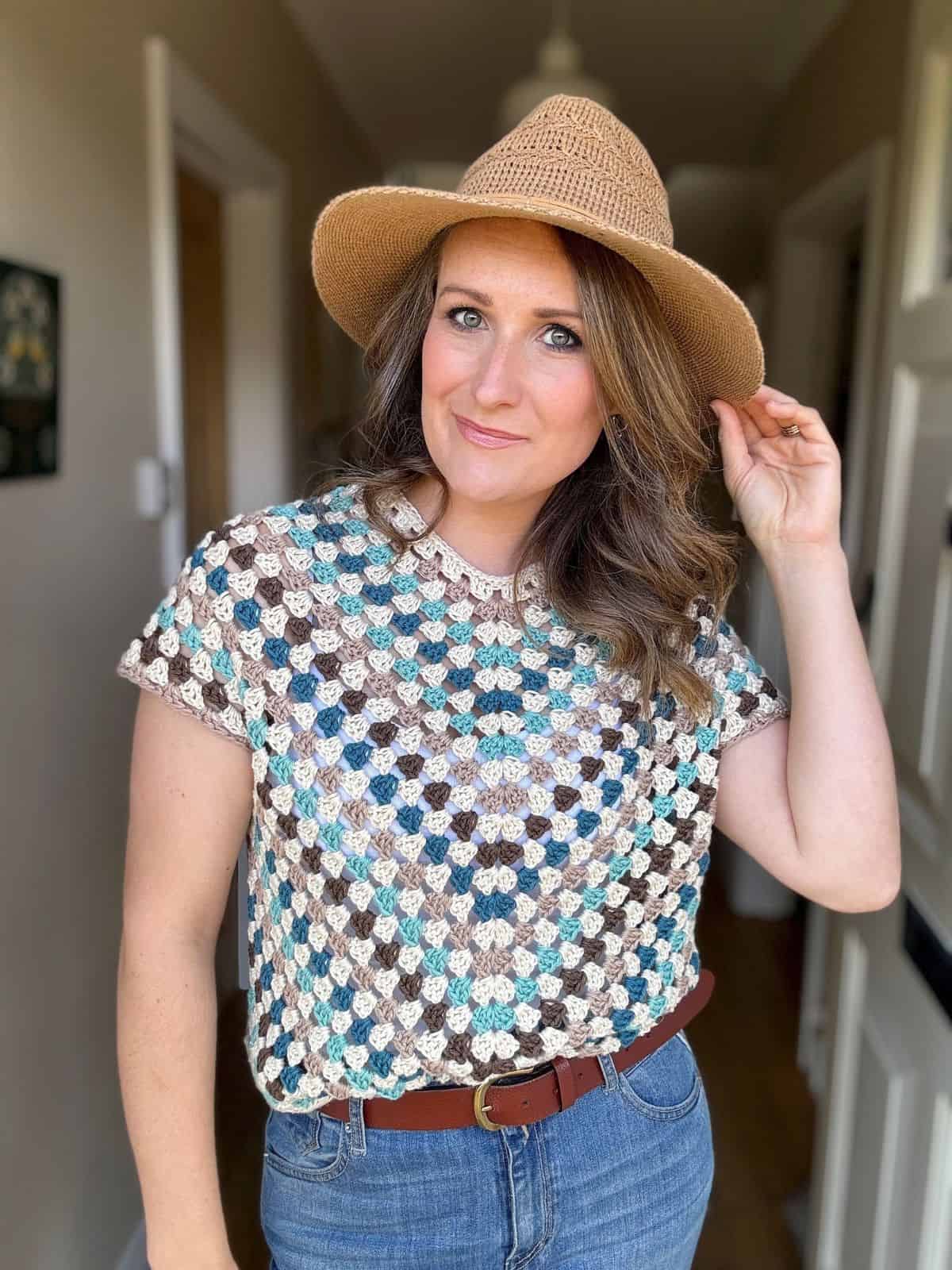 A woman is posing indoors, wearing a granny stitch crochet top, blue jeans, and a straw hat, touching the brim of the hat with her right hand. She has wavy hair, and there is a door in the background.