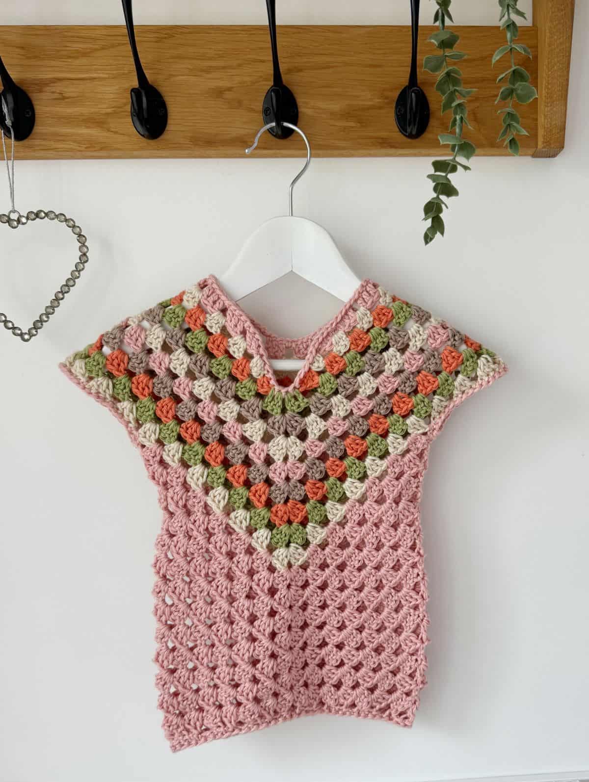 A handmade crocheted baby dress, crafted from a delightful crochet dress pattern for baby, in pink, green, and cream colors hangs on a white hanger against a wall with a wooden coat rack and a small plant.