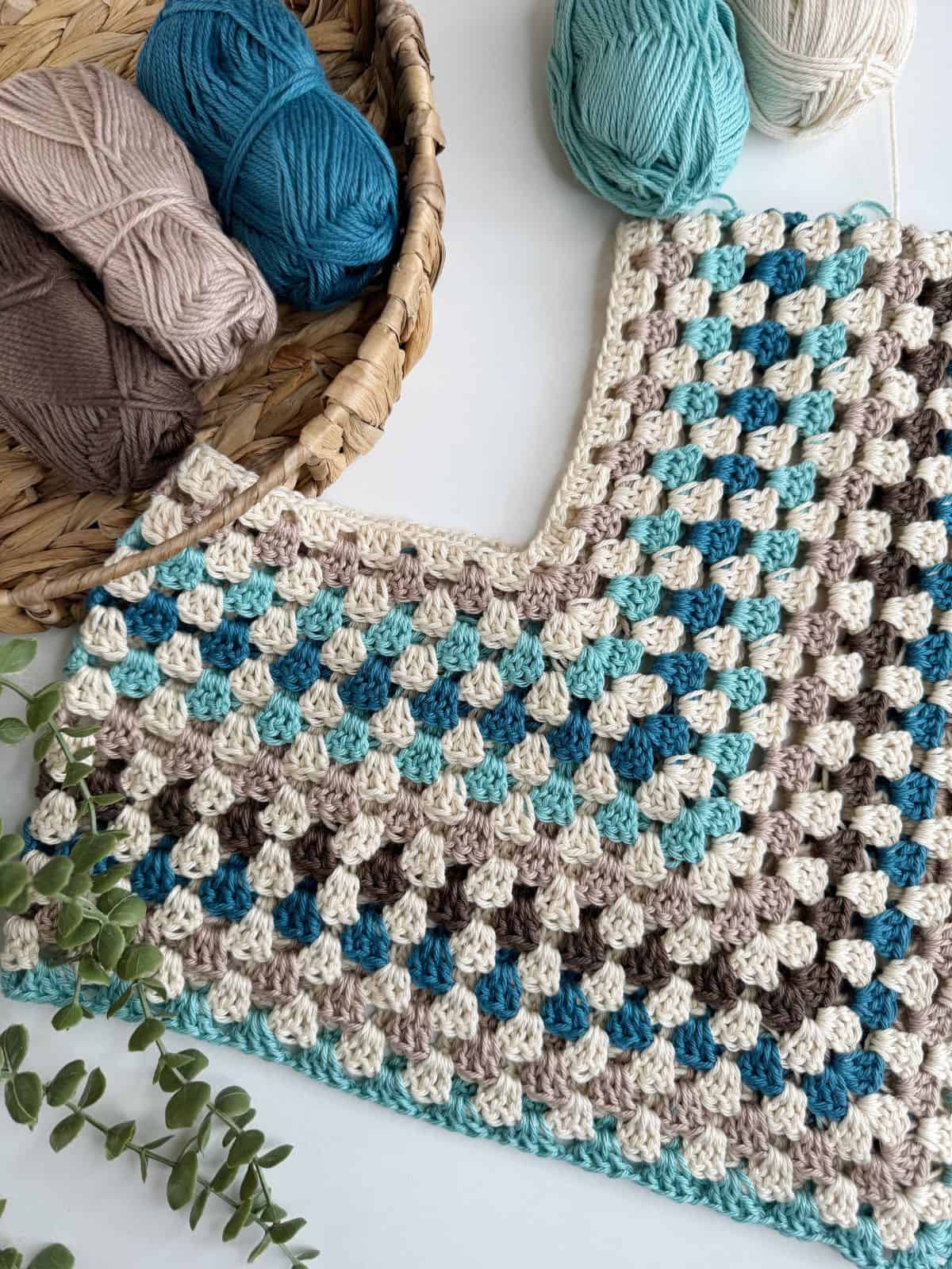 Crocheted item in progress with a colorful pattern of cream, brown, teal, and blue yarn squares. Skeins of yarn in various colors are in a wicker basket nearby, with a sprig of green leaves on the side.