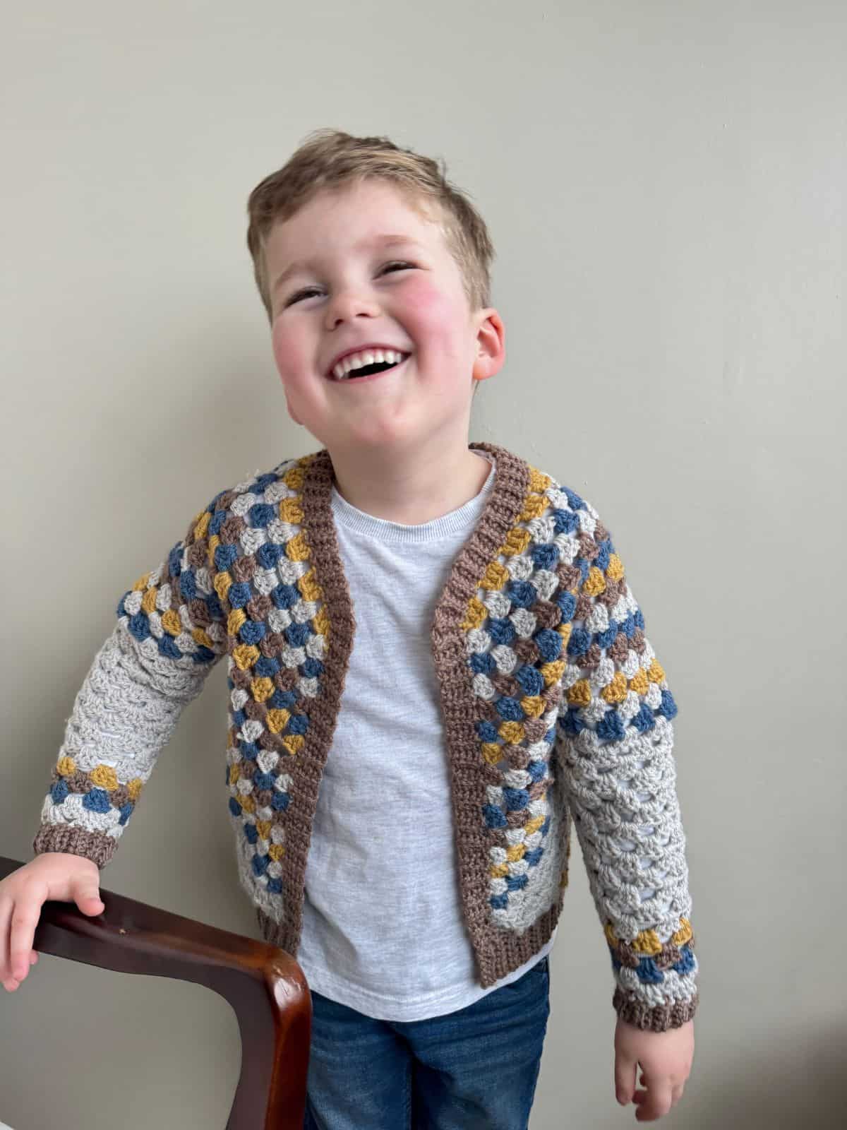 A young boy with a big smile is standing indoors, wearing a multicolored crochet cardigan over a white shirt and jeans.