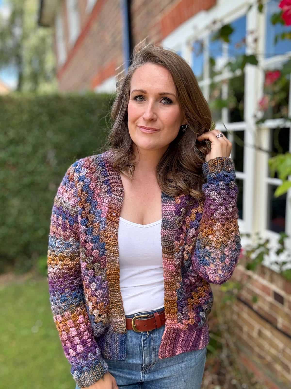 A person with long hair wearing a colorful, granny stitch crocheted cardigan, white top, and jeans stands outside in front of a brick building with greenery in the background.