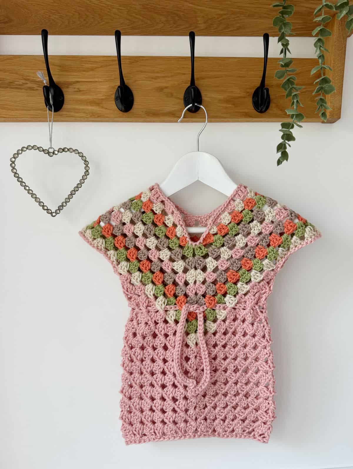 A crocheted pink baby dress made from a delightful crochet dress pattern for baby, featuring a multi-colored yoke, hangs on a white hanger against a white wall. Above it, a wooden coat rack adds charm while a heart-shaped decor and hanging plant complete the scene.