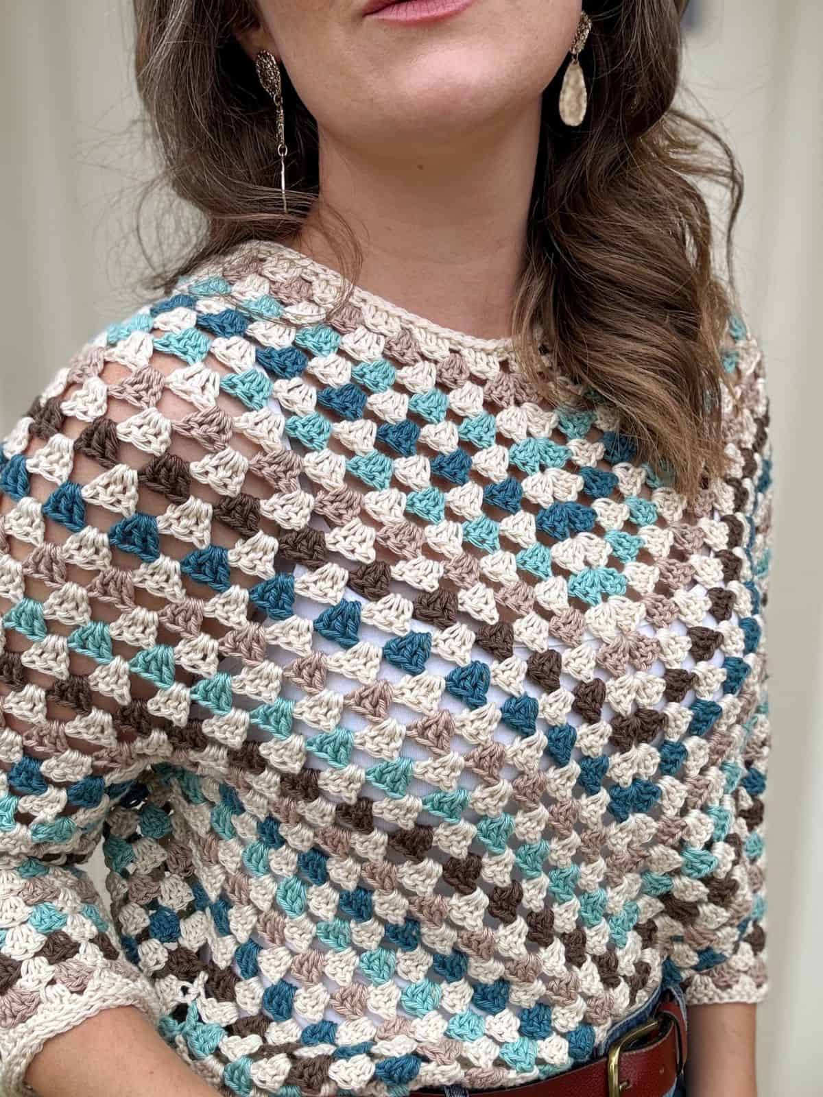 A person is wearing a colorful, crocheted top with a mix of light blue, white, brown, and beige hues, and a pair of dangling earrings. The person's face is partially out of frame.