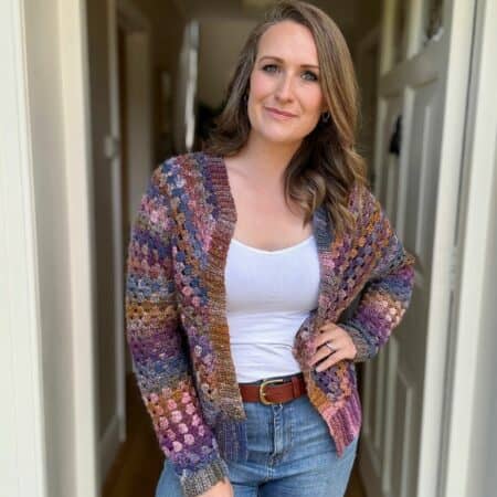 A woman stands in a hallway wearing a colorful knitted cardigan, white top, and jeans. She is looking at the camera with a slight smile, with one hand in her pocket and the other resting on her hip.