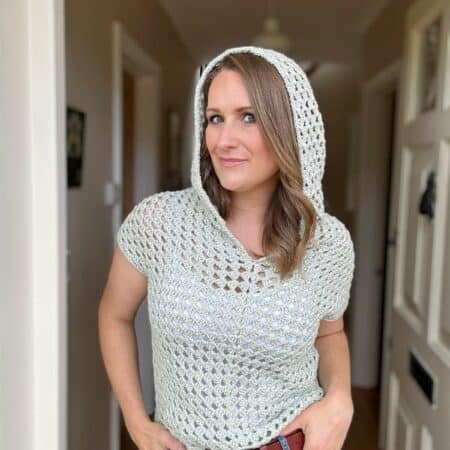 A woman stands in a hallway wearing a light knitted hooded top and jeans, with hands in her pockets.