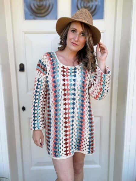 Person standing in front of a white door, wearing a colorful crocheted dress with stripes and a brown hat. They are touching the hat with one hand while looking at the camera.