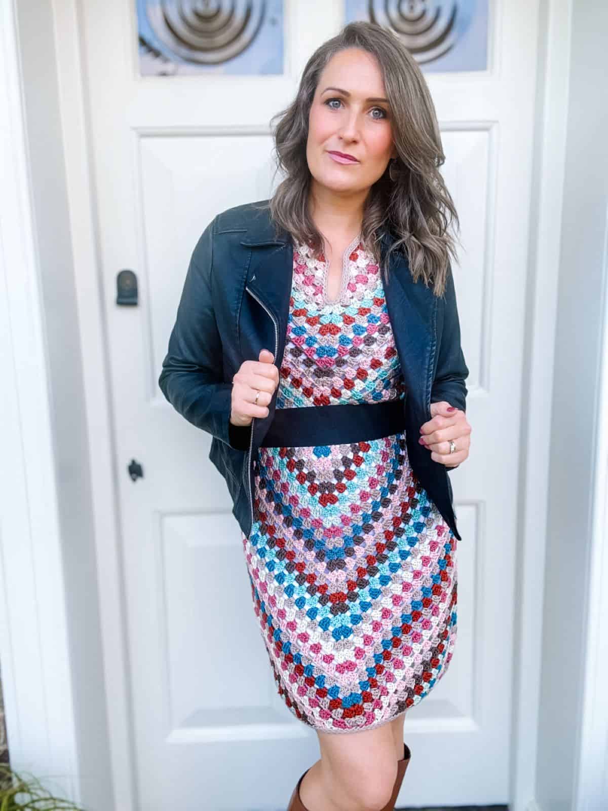 A woman with long, wavy hair stands in front of a white door, wearing a black jacket over a vibrant summer crochet dress pattern. She looks directly at the camera with a serious expression.