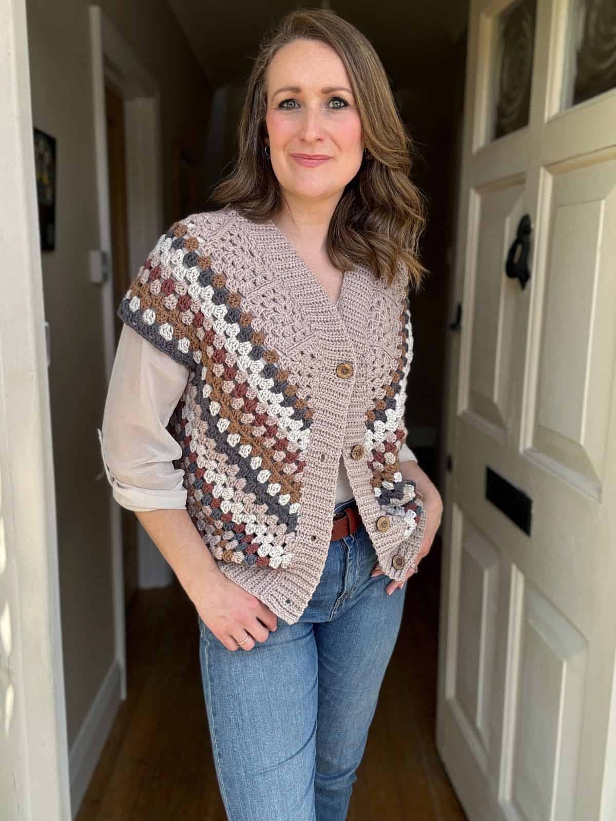 A woman stands indoors, wearing a crochet cardigan with a geometric pattern over a white blouse and blue jeans. She is looking at the camera with her hands in her pockets.