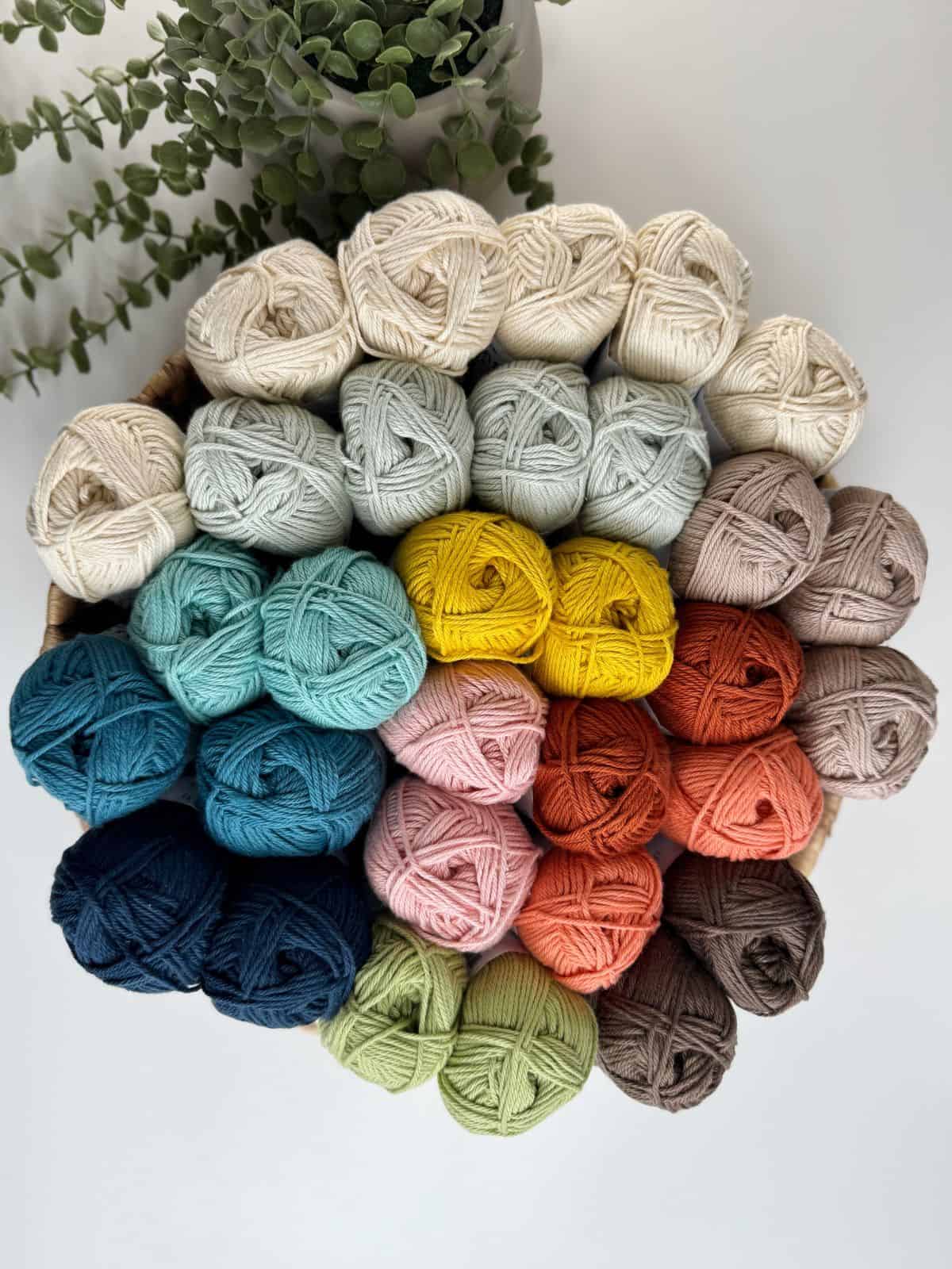 A basket filled with neatly arranged skeins of yarn in various colors including shades of white, blue, green, yellow, pink, orange, and brown. Greenery can be seen in the background.