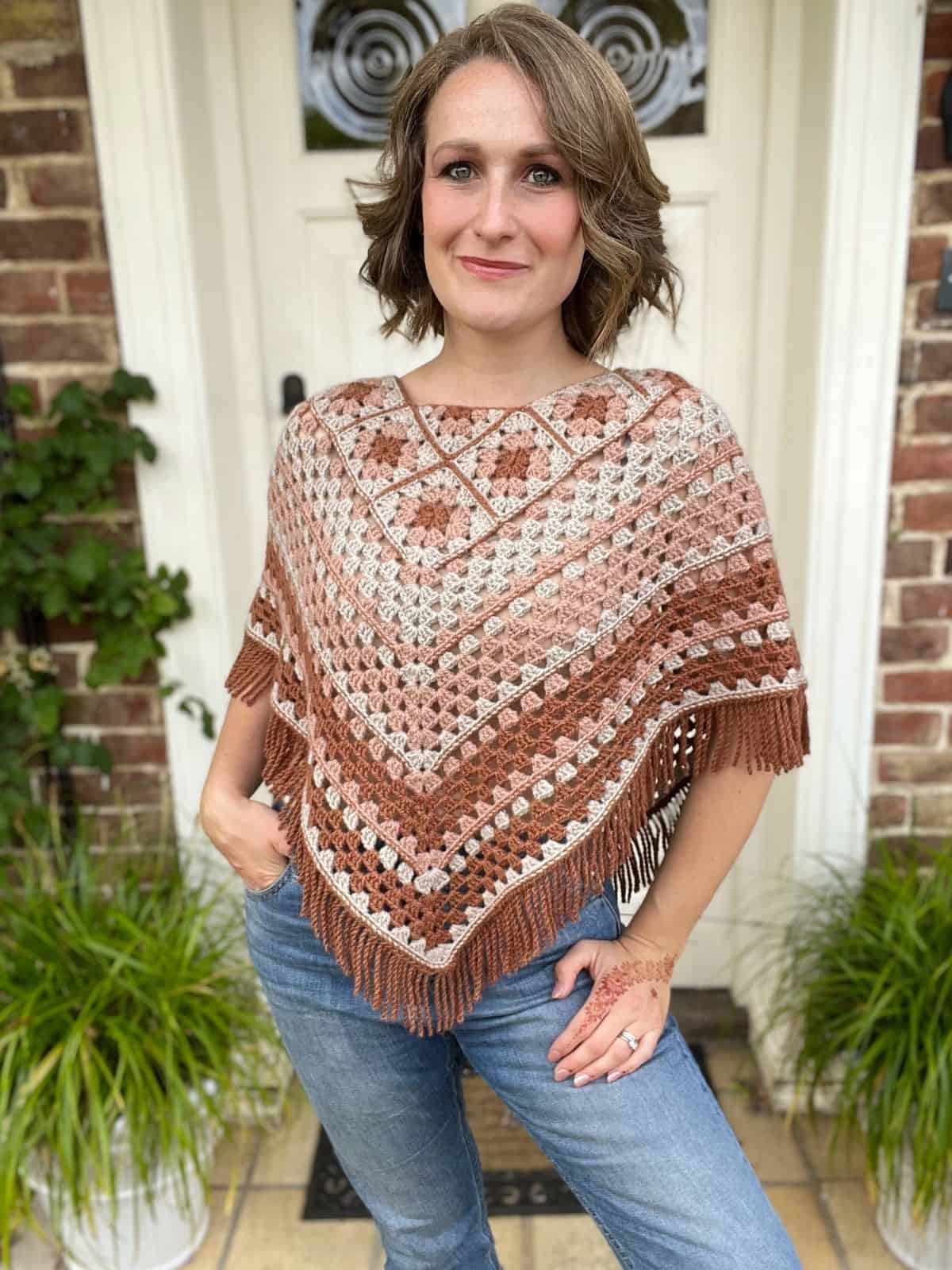 A person stands in front of a brick house entrance wearing a patterned poncho and blue jeans, with one hand resting on their hip and the other displaying a henna design. Green plants flank the doorway.