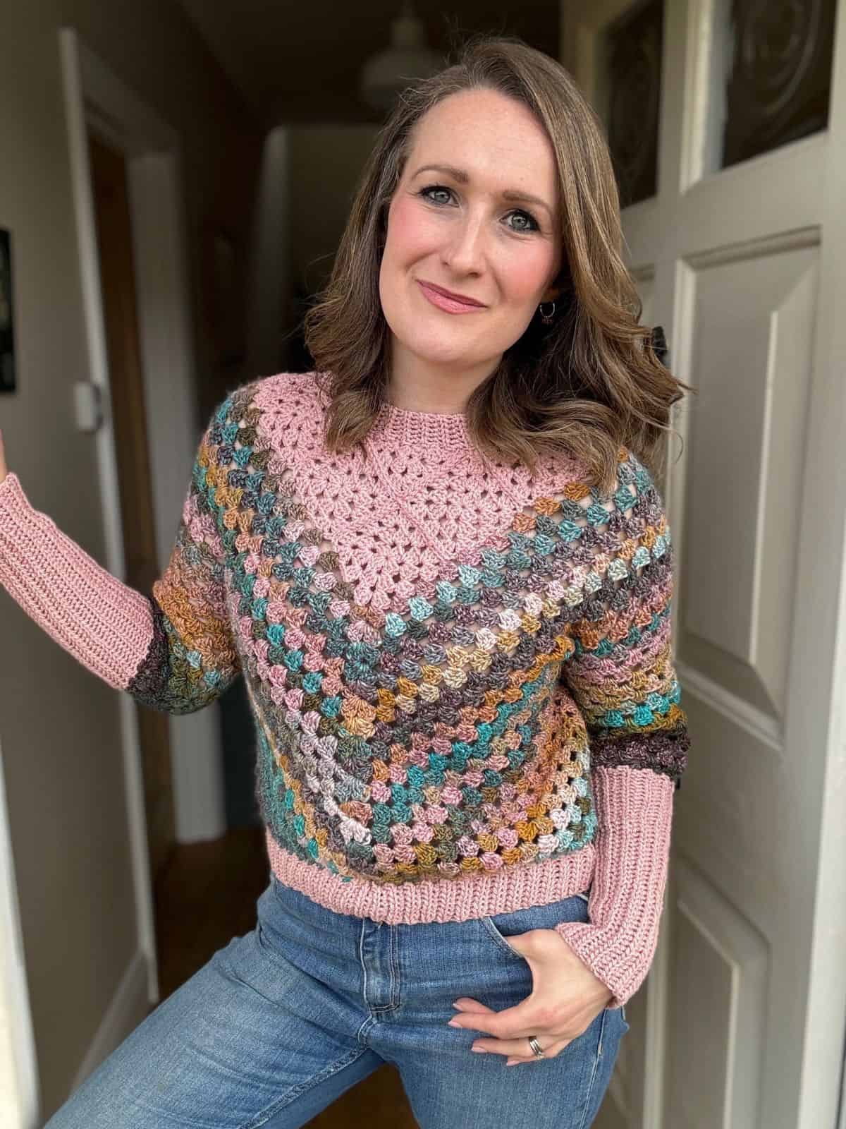 A person stands in a doorway wearing a colorful, crocheted sweater with pink, green, brown, and blue patterns, paired with blue jeans.