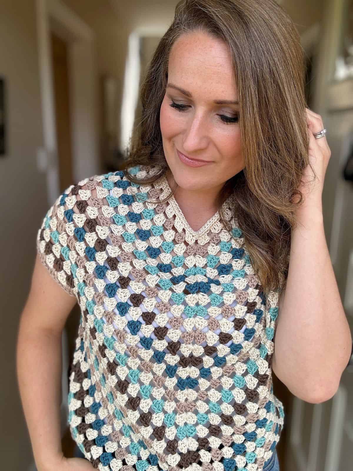 A woman with long brown hair stands indoors, wearing a crocheted top in shades of blue, brown, and white. She looks down and touches her hair with one hand.