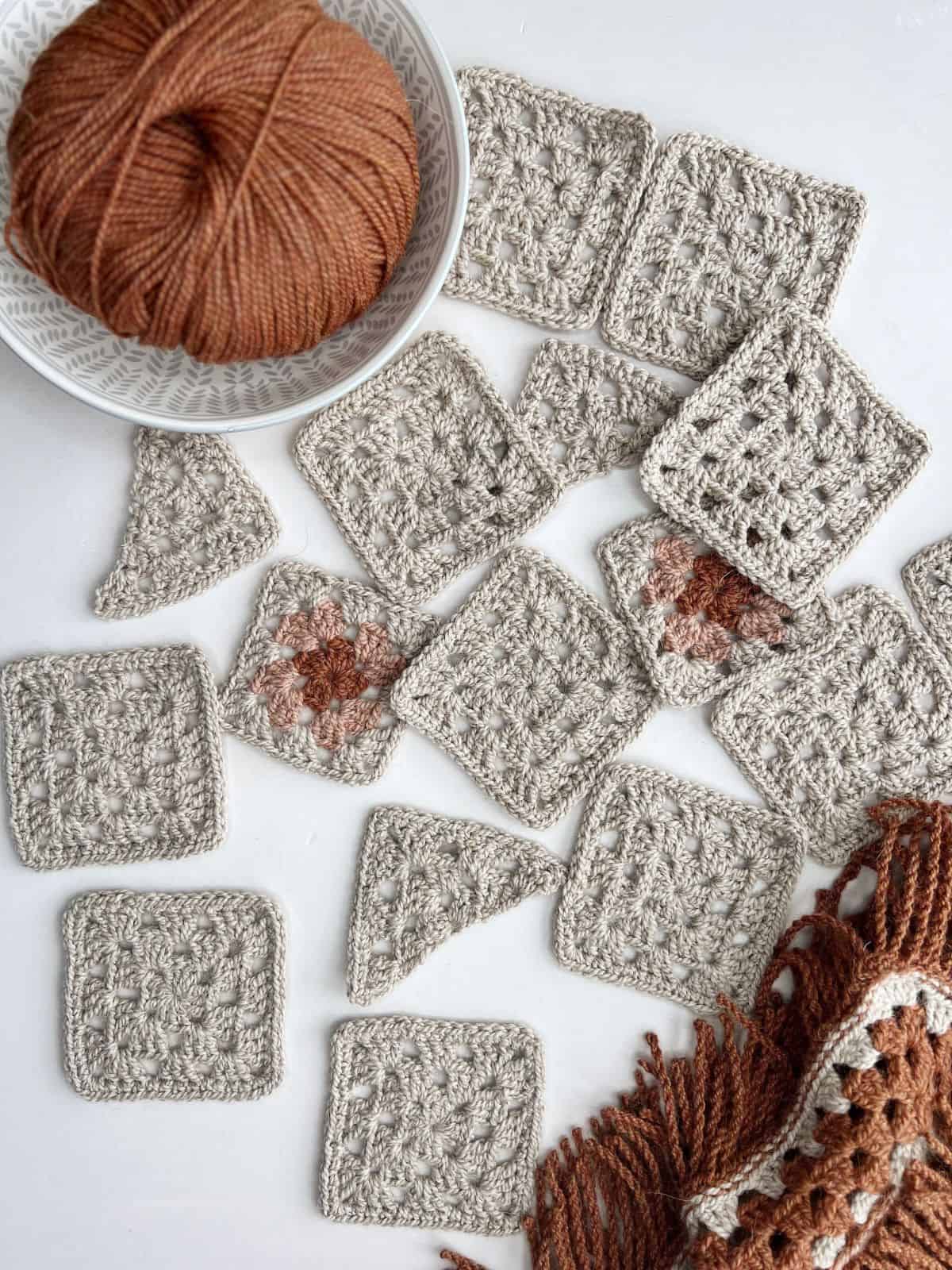 Yarn and completed gray and brown crochet squares and triangles spread out on a white surface.