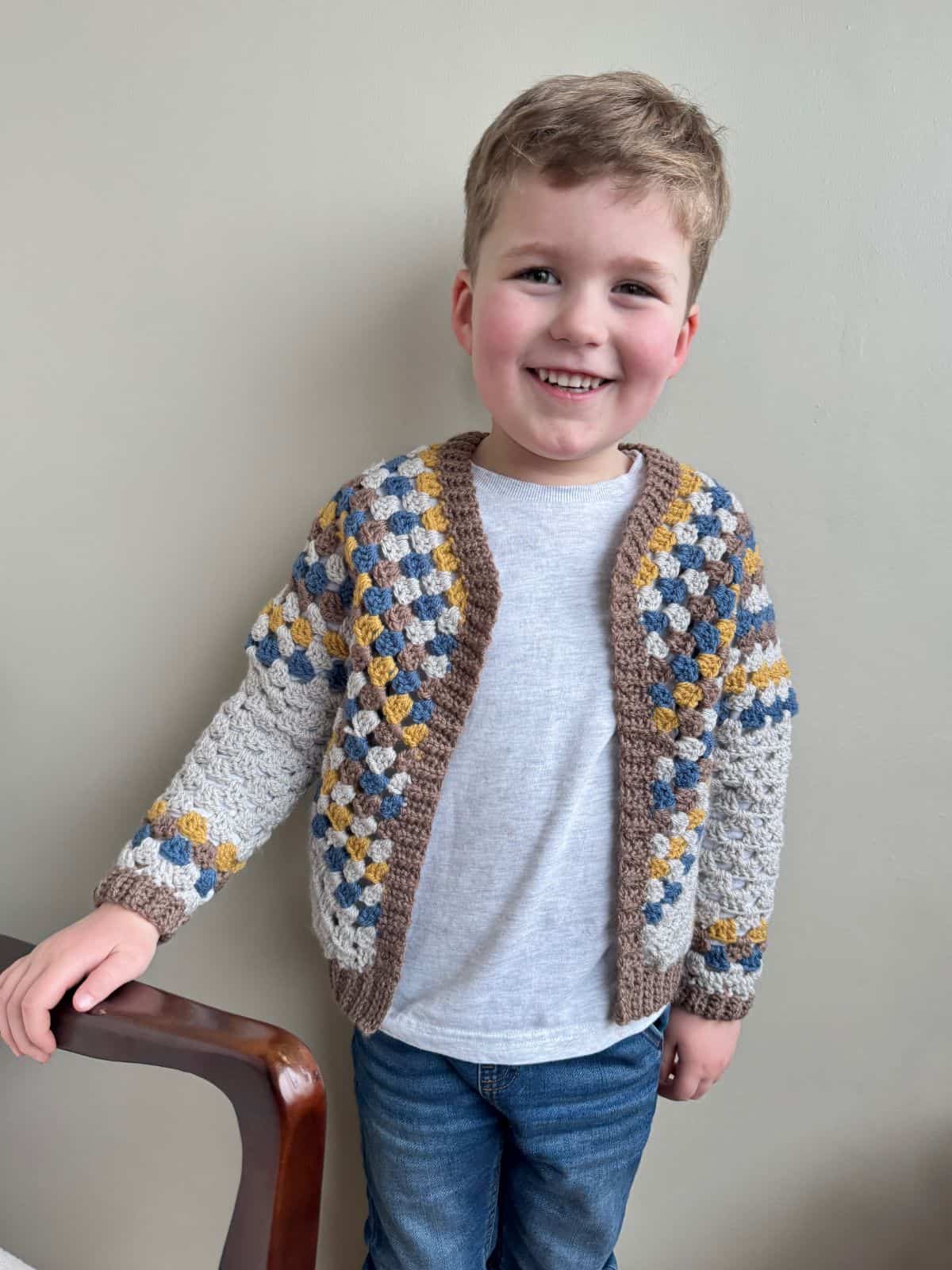 A smiling young boy in a white t-shirt and a crocheted cardigan with blue, yellow, and gray patterns stands next to a wooden chair.