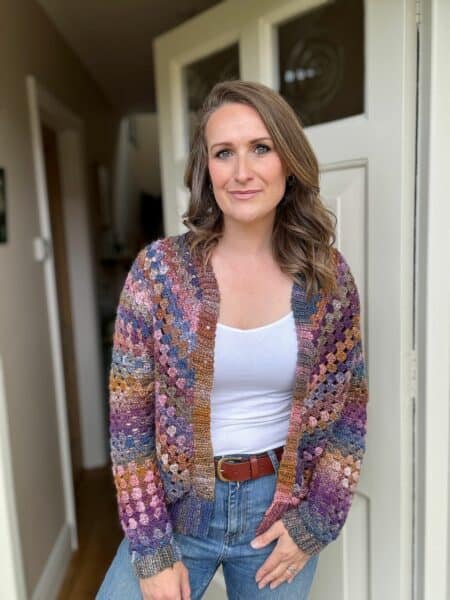 A person stands indoors wearing a colorful granny stitch cardigan over a white top and jeans, with one hand in their pocket and a neutral expression. The background includes a partially open door.