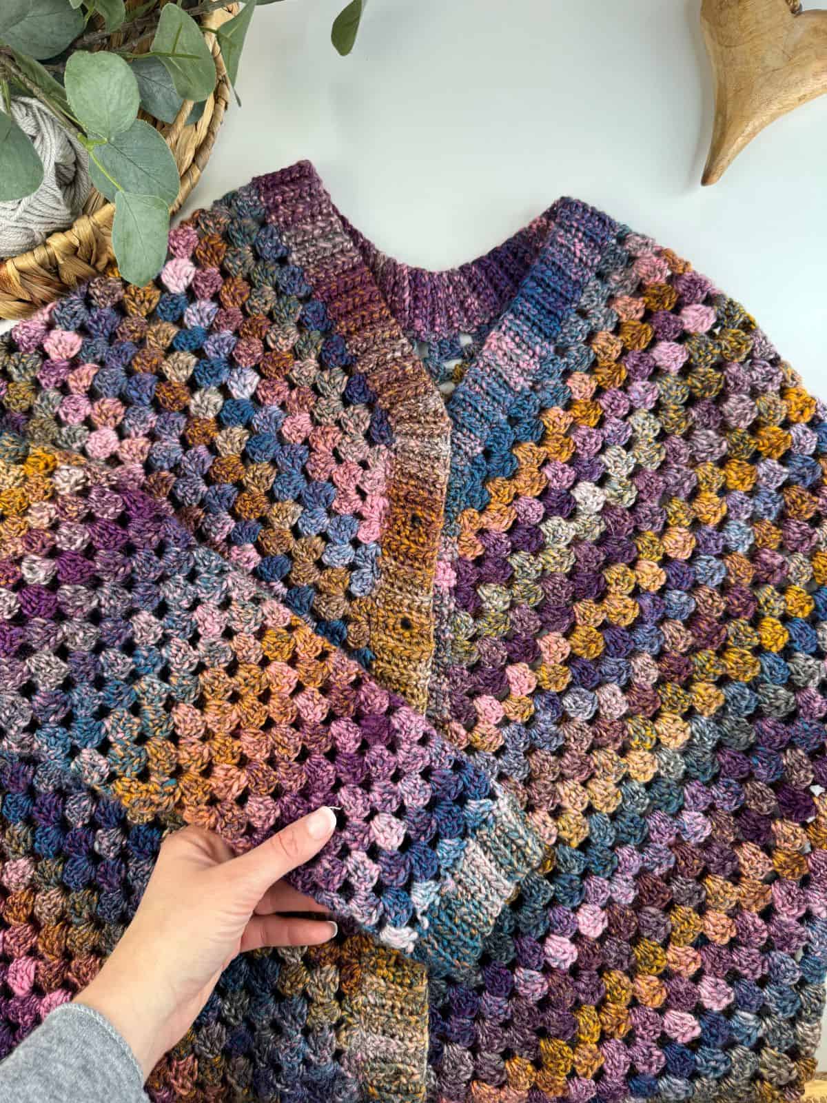 A hand holding the sleeve of a colorful crochet sweater, featuring a granny stitch cardigan pattern with squares in shades of purple, blue, orange, and pink. Green leaves and a wicker basket create a charming backdrop.