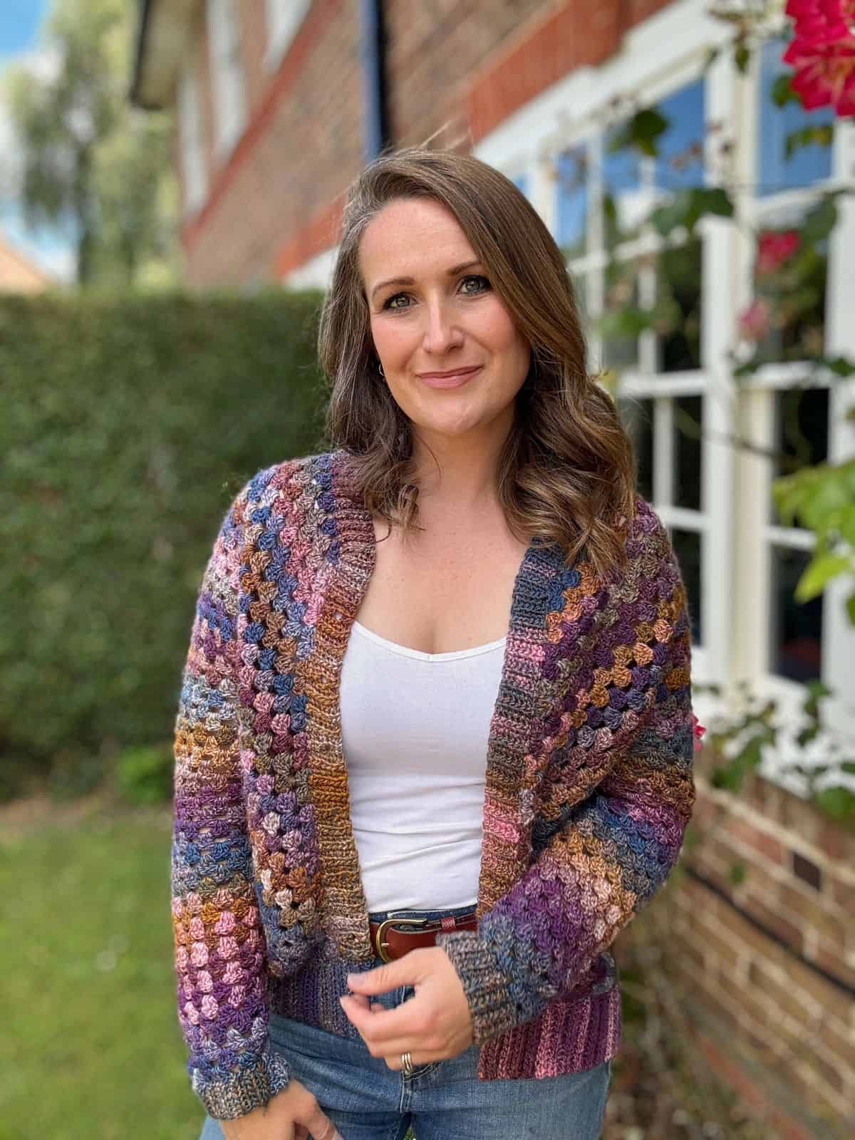 A woman stands outside in front of a brick house with a window. She is wearing a colorful crocheted cardigan, a white top, and jeans, and is smiling at the camera.