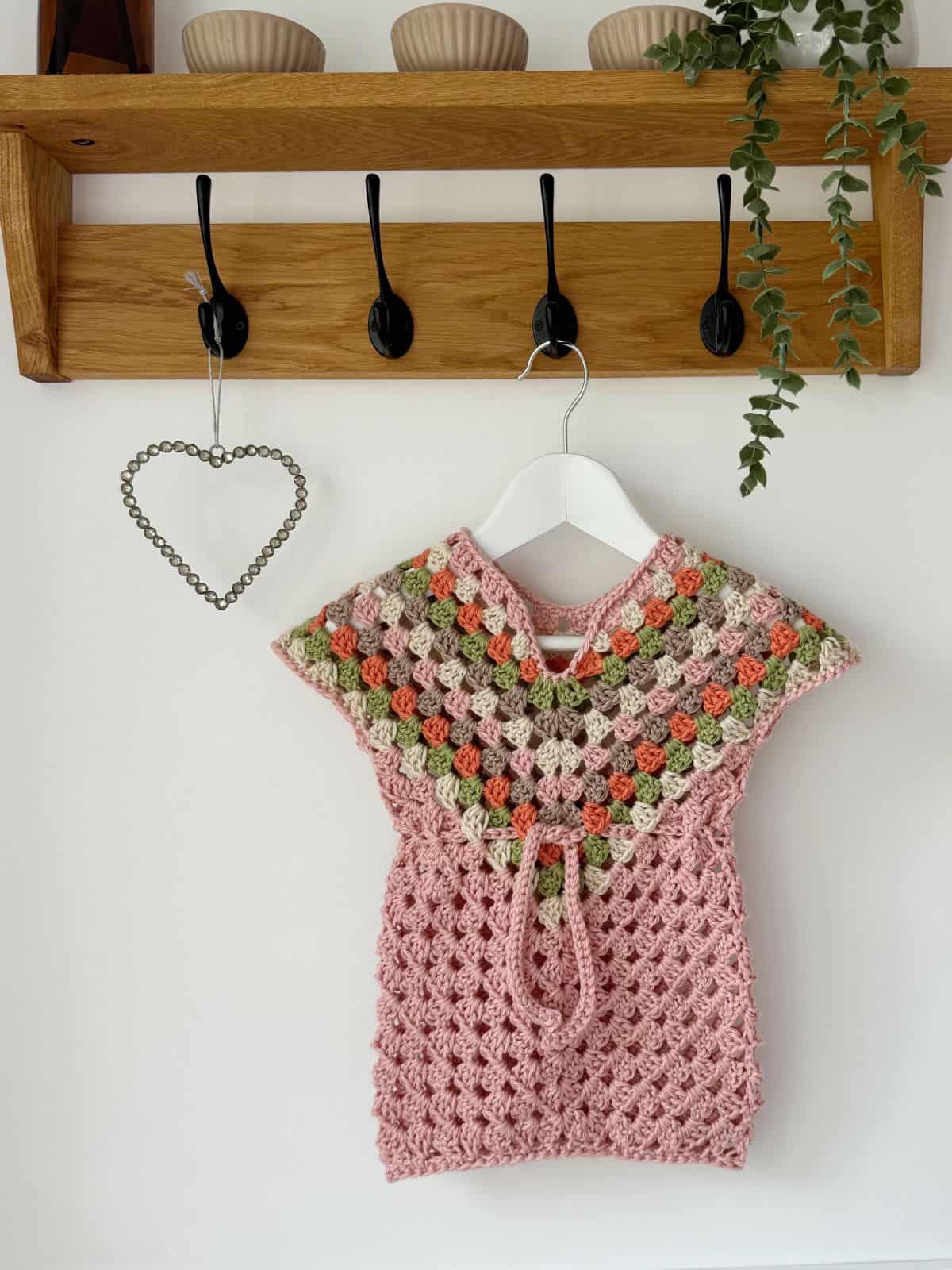 A crocheted pink and multicolored baby dress hangs on a white hanger from a wooden coat rack with five black hooks, next to a hanging green plant and a metal heart decor.