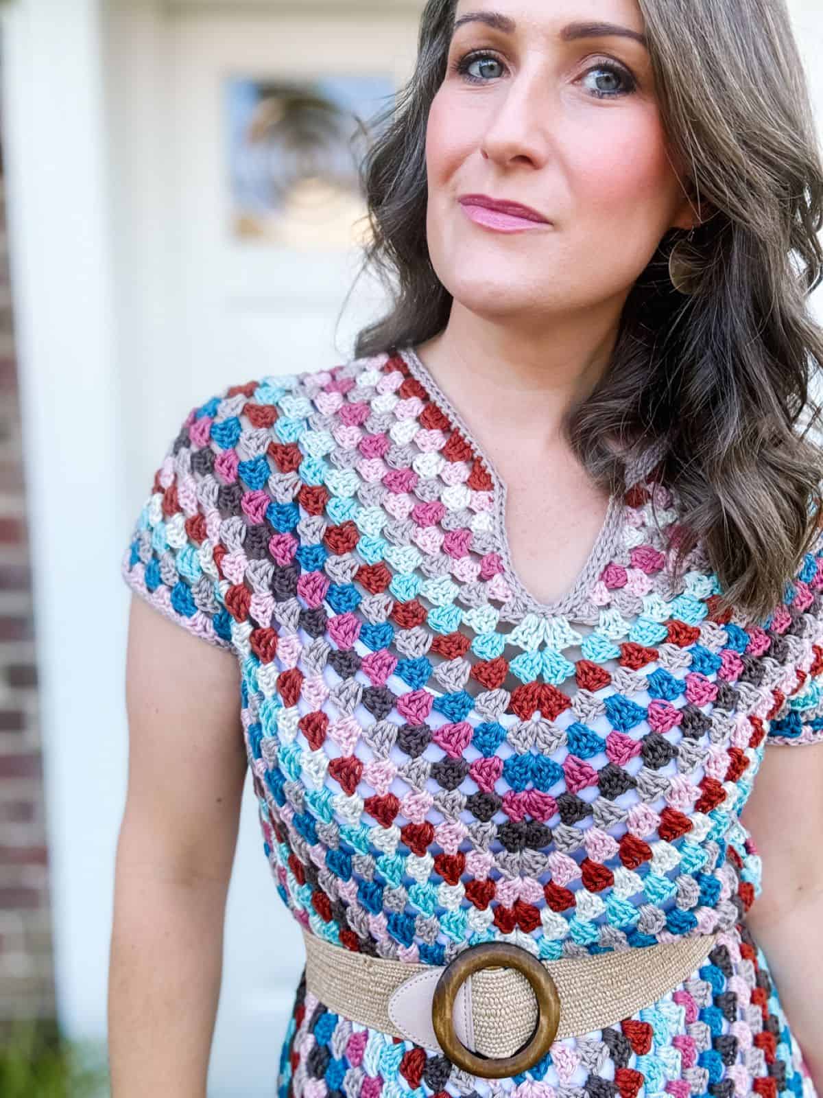 A person with wavy hair wearing a colorful crochet dress featuring a summer crochet dress pattern and a beige belt with a large circular buckle stands in front of a blurred background.