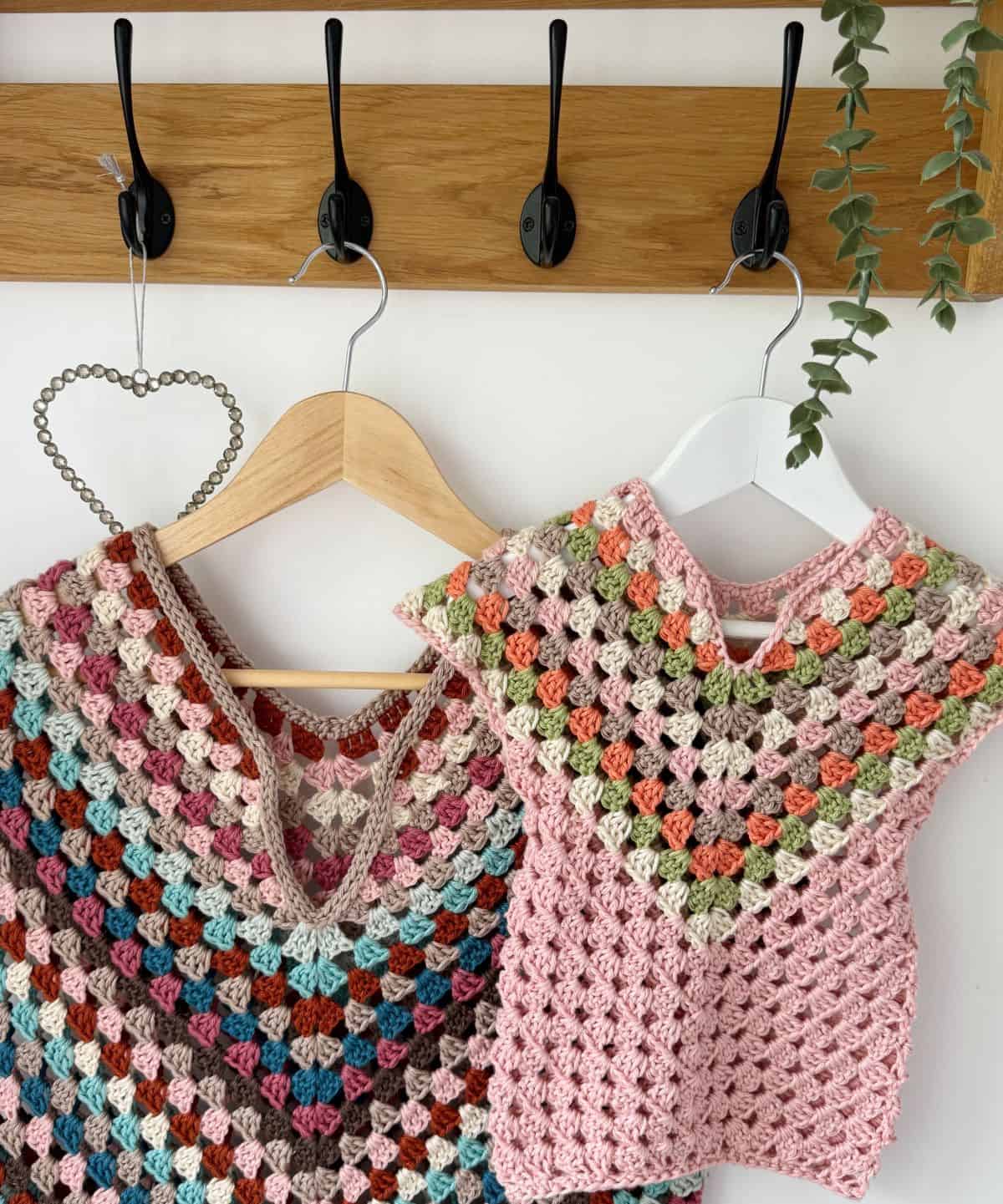 Two crocheted dresses on white and wooden hangers are displayed on a wooden wall hook. One dress is multi-colored and adult-sized while the other is pink and child-sized, showcasing an adorable crochet dress pattern for baby. A small green plant is in the background.