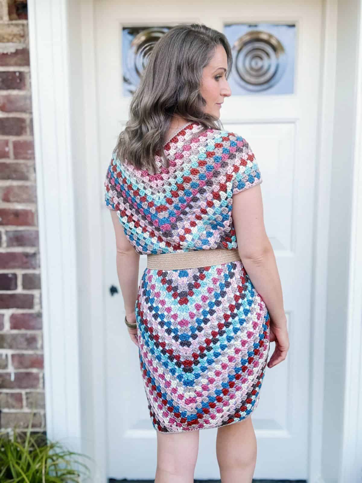 A woman with long, wavy hair stands with her back to the camera, wearing a colorful crocheted dress with a pattern of diagonal lines, in front of a white door and a brick wall.