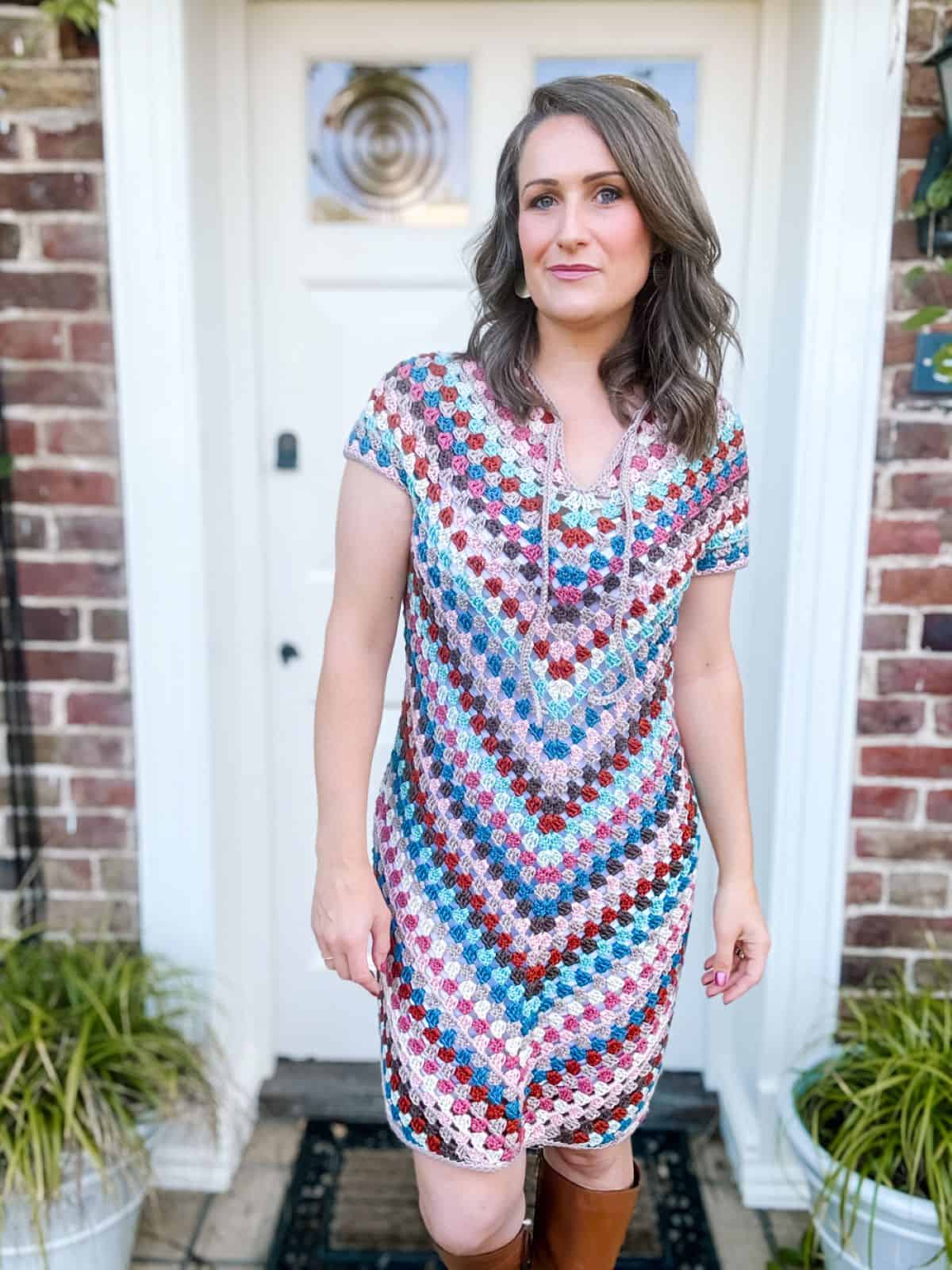 A woman stands in front of a doorway, wearing a colorful summer crochet dress pattern and brown boots. She has shoulder-length hair and a neutral expression. Plants are placed on both sides of the entrance.