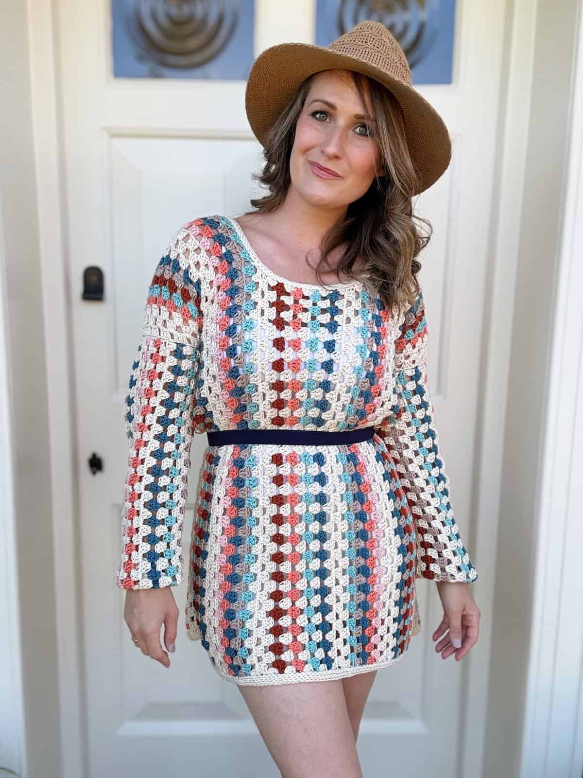 A person stands in front of a door, wearing a woven, multicolored crochet granny dress and a brown hat.
