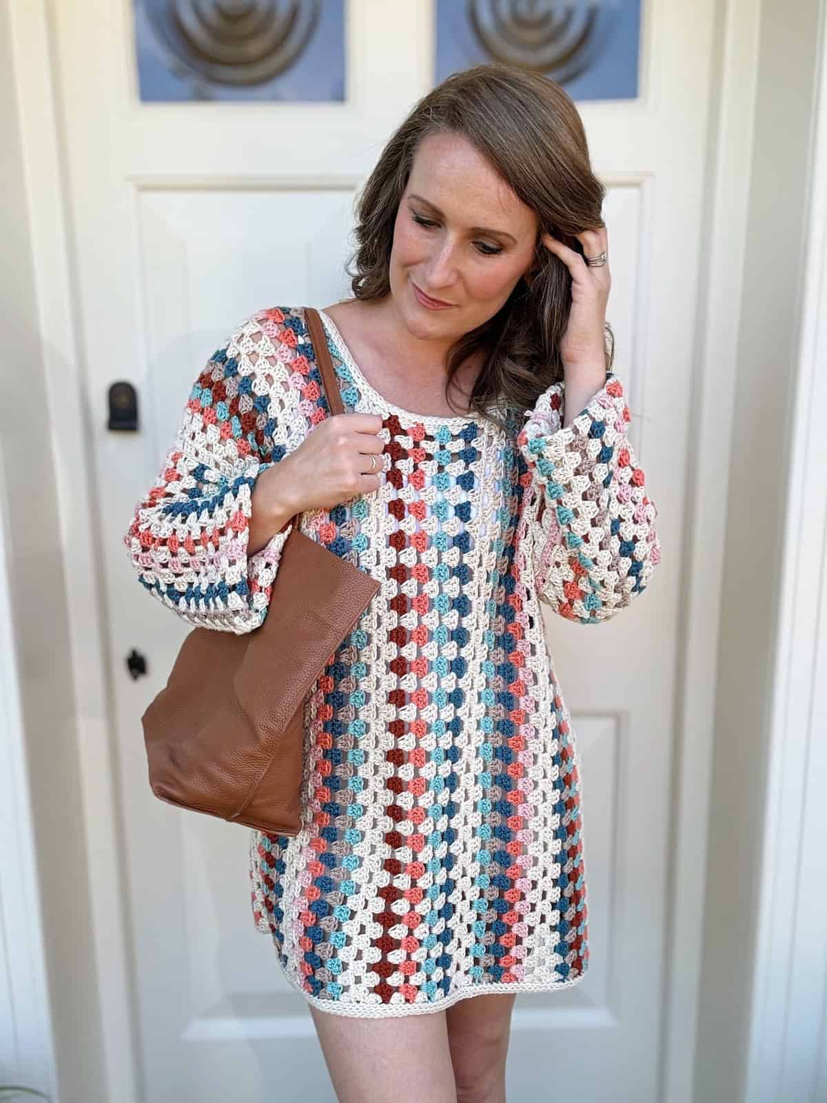 A person stands in front of a white door, wearing a colorful, crochet sweater dress and holding a brown leather bag over their shoulder.